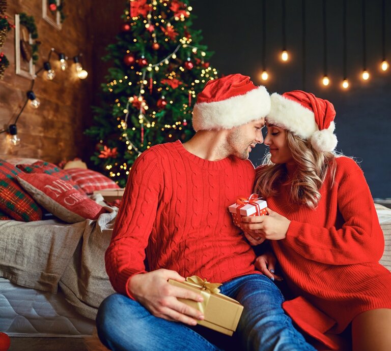 Ein verliebtes Paar in roten Weihnachtsoutfits und Weihnachtsmützen sitzt vor einem festlich geschmückten Weihnachtsbaum. Sie halten sich gegenseitig Geschenke und lächeln sich zärtlich an, während im Hintergrund eine gemütliche, weihnachtliche Szenerie mit Lichterketten und dekorierten Kissen zu sehen ist. | © studioroman via Canva.com