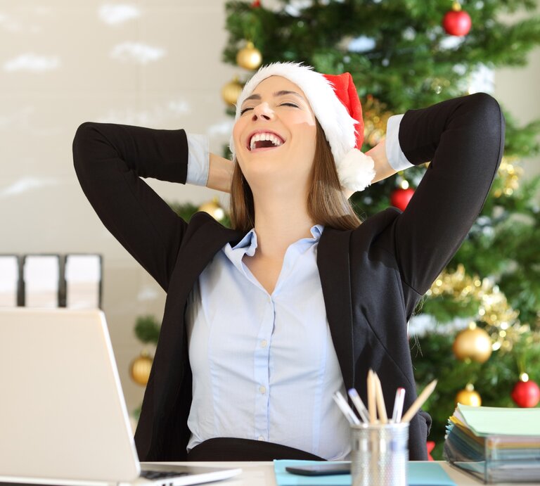 Eine lachende Frau in einem Büro, die eine Weihnachtsmütze trägt, sitzt entspannt zurückgelehnt vor ihrem Laptop. Im Hintergrund steht ein geschmückter Weihnachtsbaum mit goldenen und roten Kugeln. | © Getty Images/ AntonioGuillem via Canva.com