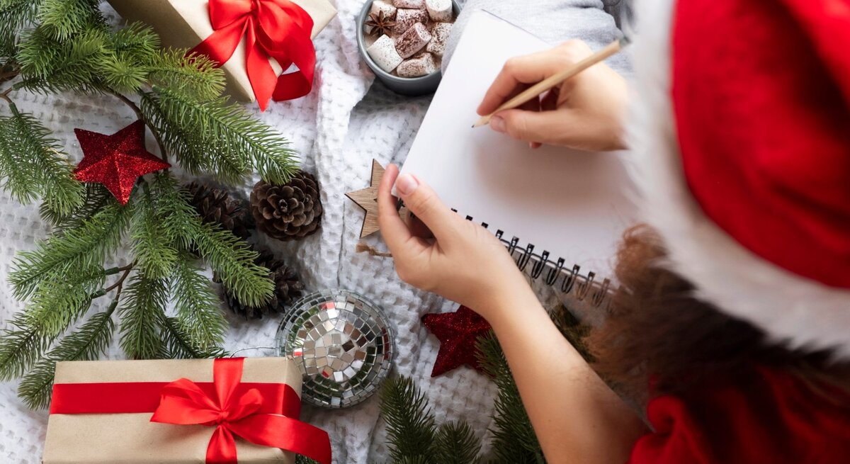 Ein weihnachtliches Bild zeigt eine Person, die in ein leeres Notizbuch schreibt. Sie trägt eine rote Weihnachtsmütze und sitzt auf einer kuscheligen, weißen Decke. Um die Person herum liegen festlich verpackte Geschenke mit roten Schleifen, grüne Tannenzweige, rote Weihnachtssterne, Tannenzapfen und eine glänzende silberne Christbaumkugel. In der Nähe steht eine Tasse heiße Schokolade mit Marshmallows, was eine gemütliche und festliche Atmosphäre schafft.