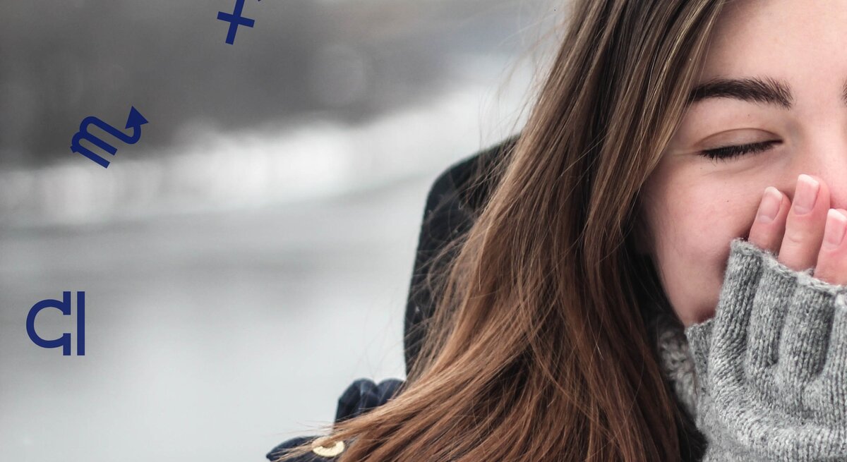 Eine junge Frau mit langen braunen Haaren, die eine dunkle Jacke und graue Wollhandschuhe trägt. Sie hält lächelnd ihre Hände an ihr Gesicht, während sie die Augen geschlossen hat. Im Hintergrund ist eine winterliche Landschaft mit verschwommenem Wasser und grauem Himmel zu sehen. Um sie herum schweben Symbole der Sternzeichen, wie Schütze und Skorpion.