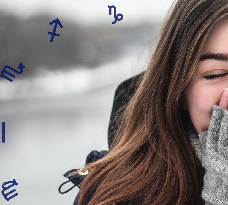 Eine junge Frau mit langen braunen Haaren, die eine dunkle Jacke und graue Wollhandschuhe trägt. Sie hält lächelnd ihre Hände an ihr Gesicht, während sie die Augen geschlossen hat. Im Hintergrund ist eine winterliche Landschaft mit verschwommenem Wasser und grauem Himmel zu sehen. Um sie herum schweben Symbole der Sternzeichen, wie Schütze und Skorpion. | © Pexels/ pixabay und Chikovnaya via Canva.com [M]