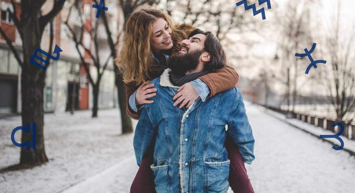 Ein glückliches Paar genießt einen winterlichen Spaziergang: Die Frau trägt eine braune Jacke und umarmt den Mann lachend von hinten, während sie auf seinem Rücken sitzt. Der Mann, in einer Jeansjacke mit Schafsfellkragen, lächelt herzlich. Im Hintergrund sind verschneite Bäume und eine städtische Szenerie zu sehen. Um das Paar herum schweben astrologische Symbole in Blau, die eine Verbindung zur Astrologie herstellen. Das Bild vermittelt Freude, Liebe und die Harmonie zwischen den Sternzeichen.
