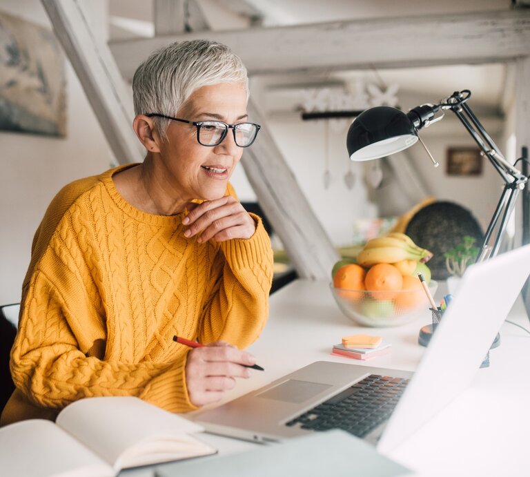 Eine ältere Frau mit grauem, kurzem Haar und Brille sitzt an einem Schreibtisch und arbeitet konzentriert an ihrem Laptop. Sie trägt einen gemütlichen gelben Strickpullover und hält einen Stift in der Hand. Auf dem Schreibtisch liegen ein aufgeschlagenes Notizbuch, Post-its und eine Schreibtischlampe. Im Hintergrund stehen eine Schale mit frischem Obst und dezente Einrichtungsdetails, die eine helle und freundliche Arbeitsatmosphäre schaffen. | ©  Getty Images Signature/ RgStudio via Canva.com