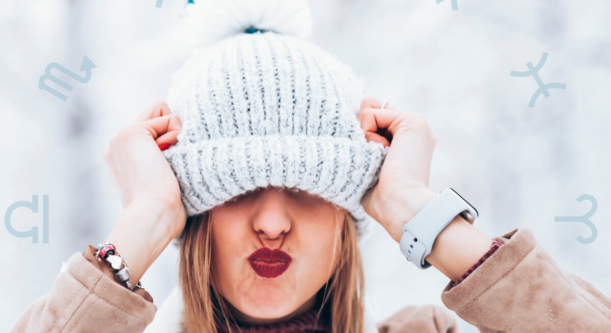Eine fröhliche Frau in winterlicher Kleidung zieht ihre gestrickte Bommelmütze spielerisch über die Augen und macht einen Kussmund. Der Hintergrund zeigt eine verschneite, helle Szenerie, und astrologische Symbole sind dezent über das Bild verstreut. Das Bild vermittelt Lebensfreude, Verspieltheit und einen Hauch von Astrologie.