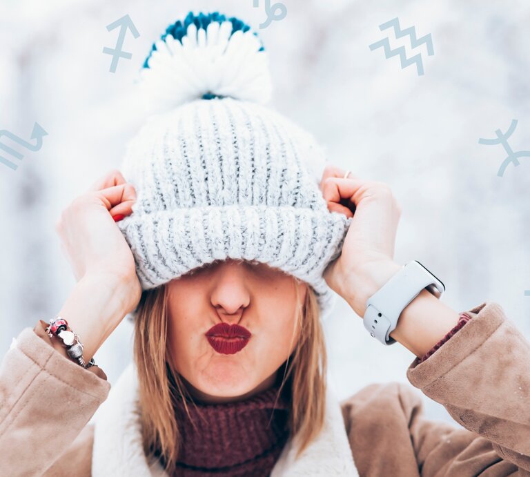 Eine fröhliche Frau in winterlicher Kleidung zieht ihre gestrickte Bommelmütze spielerisch über die Augen und macht einen Kussmund. Der Hintergrund zeigt eine verschneite, helle Szenerie, und astrologische Symbole sind dezent über das Bild verstreut. Das Bild vermittelt Lebensfreude, Verspieltheit und einen Hauch von Astrologie. | © Kseniia Perminova und Chikovnaya via Canva.com [M]