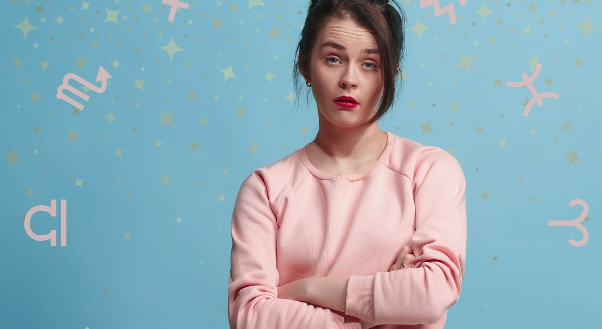 Eine Frau in einem rosa Pullover steht vor einem hellblauen Hintergrund mit pastellfarbenen Sternzeichen-Symbolen und goldenen Sternen. Sie hat die Arme verschränkt und einen nachdenklichen Gesichtsausdruck, der Fragen oder Unsicherheiten ausdrücken könnte. Das Bild symbolisiert astrologische Themen und persönliche Reflektion im Zusammenhang mit den Sternzeichen.