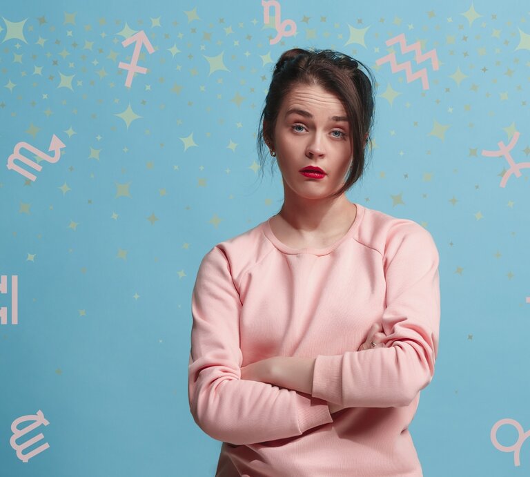 Eine Frau in einem rosa Pullover steht vor einem hellblauen Hintergrund mit pastellfarbenen Sternzeichen-Symbolen und goldenen Sternen. Sie hat die Arme verschränkt und einen nachdenklichen Gesichtsausdruck, der Fragen oder Unsicherheiten ausdrücken könnte. Das Bild symbolisiert astrologische Themen und persönliche Reflektion im Zusammenhang mit den Sternzeichen. | © Adobe Stock/ master1305, Golden shooting stars, Line gold stardust und Chikovnaya via Canva.com [M]