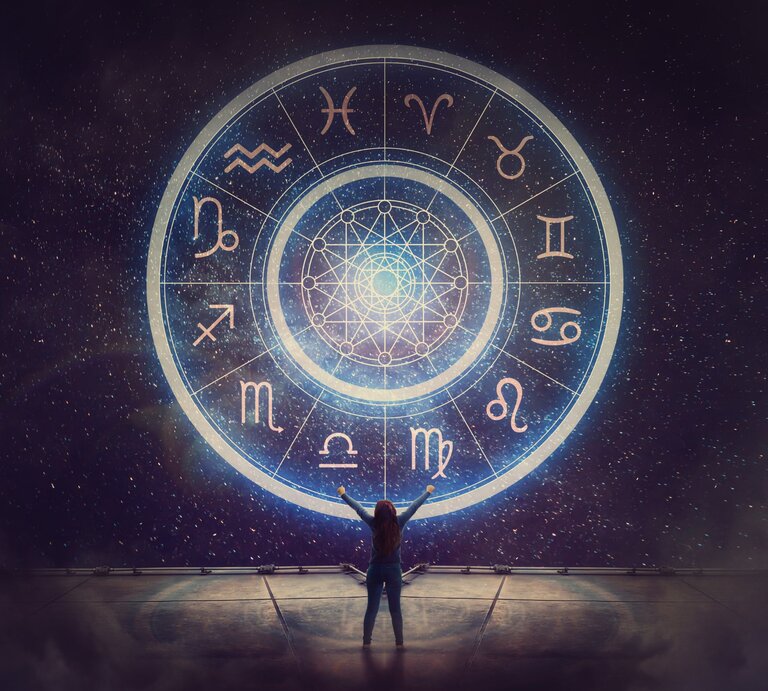 Eine Person steht vor einem großen, leuchtenden astrologischen Rad, das alle zwölf Tierkreiszeichen zeigt. Der Nachthimmel im Hintergrund ist voller funkelnder Sterne, und die Szene vermittelt eine mystische und kosmische Atmosphäre. | © Getty Images/ Bulat Silvia via Canva.com 