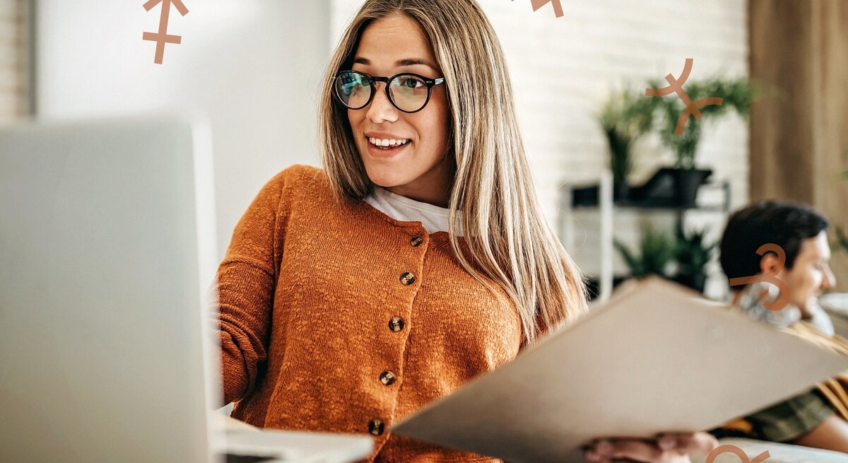 Eine Frau mit Brille und einem orangefarbenen Pullover sitzt an einem Laptop und hält einen Ordner in der Hand. Sie lächelt und wirkt konzentriert, während um sie herum astrologische Symbole schweben. Die Szene vermittelt eine produktive und motivierte Arbeitsatmosphäre mit einem Hauch von astrologischem Einfluss.