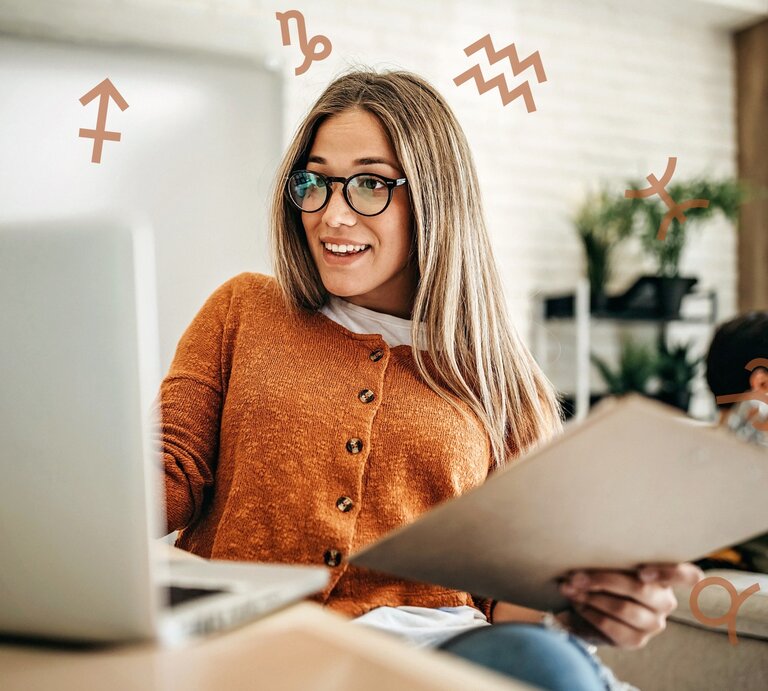 Eine Frau mit Brille und einem orangefarbenen Pullover sitzt an einem Laptop und hält einen Ordner in der Hand. Sie lächelt und wirkt konzentriert, während um sie herum astrologische Symbole schweben. Die Szene vermittelt eine produktive und motivierte Arbeitsatmosphäre mit einem Hauch von astrologischem Einfluss. | © Getty Images Signature/ eclipse_images und Chikovnaya via Canva.com [M] 