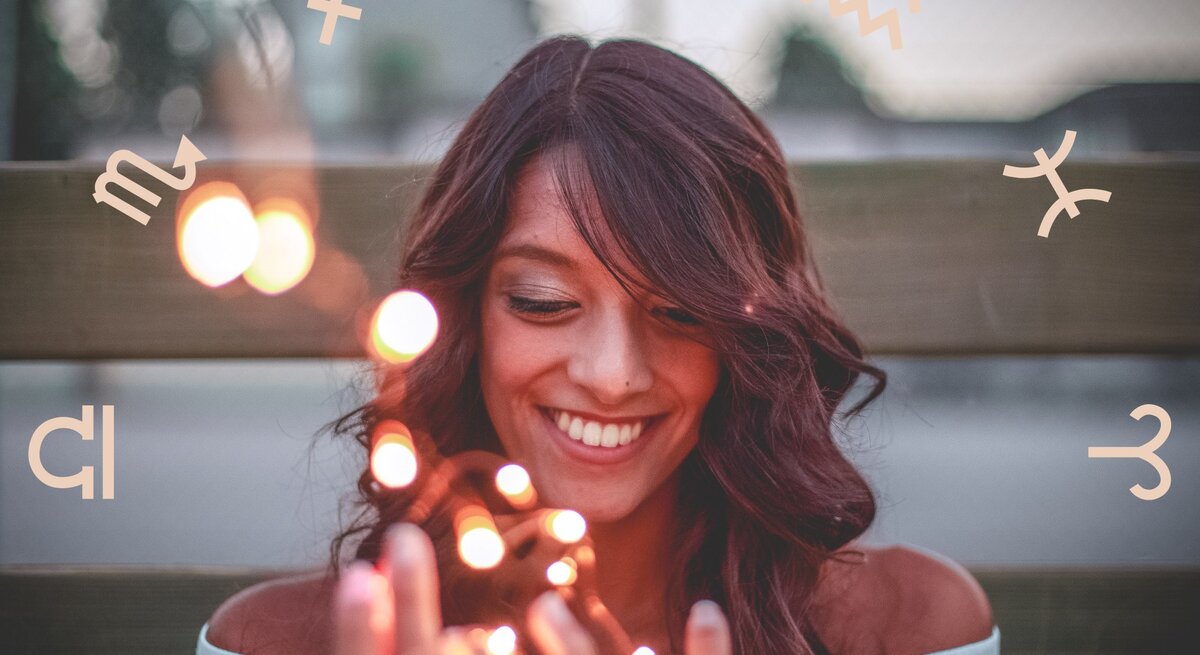 Eine lächelnde Frau mit dunklen Haaren hält leuchtende Lichter in den Händen, während um sie herum astrologische Symbole der Tierkreiszeichen schweben. Sie wirkt glücklich und entspannt, was eine positive und magische Atmosphäre vermittelt.