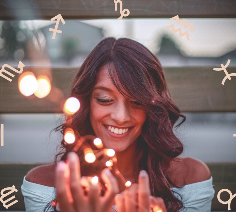 Eine lächelnde Frau mit dunklen Haaren hält leuchtende Lichter in den Händen, während um sie herum astrologische Symbole der Tierkreiszeichen schweben. Sie wirkt glücklich und entspannt, was eine positive und magische Atmosphäre vermittelt. | © Pexels/ Oleg Magni und Chikovnaya via Canva.com [M]