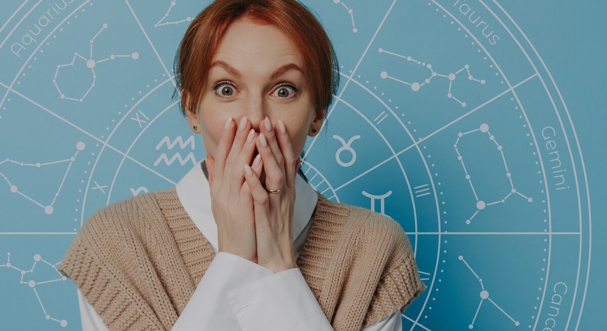 Eine überraschte Frau mit rotblonden Haaren hält sich die Hände vor den Mund und schaut mit großen Augen. Hinter ihr ist ein blauer Hintergrund mit einem astrologischen Rad und den Symbolen der Tierkreiszeichen zu sehen.