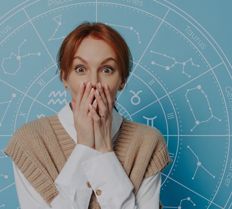 Eine überraschte Frau mit rotblonden Haaren hält sich die Hände vor den Mund und schaut mit großen Augen. Hinter ihr ist ein blauer Hintergrund mit einem astrologischen Rad und den Symbolen der Tierkreiszeichen zu sehen. | © WinWin.artlab und vkstudio via Canva.com [M]
