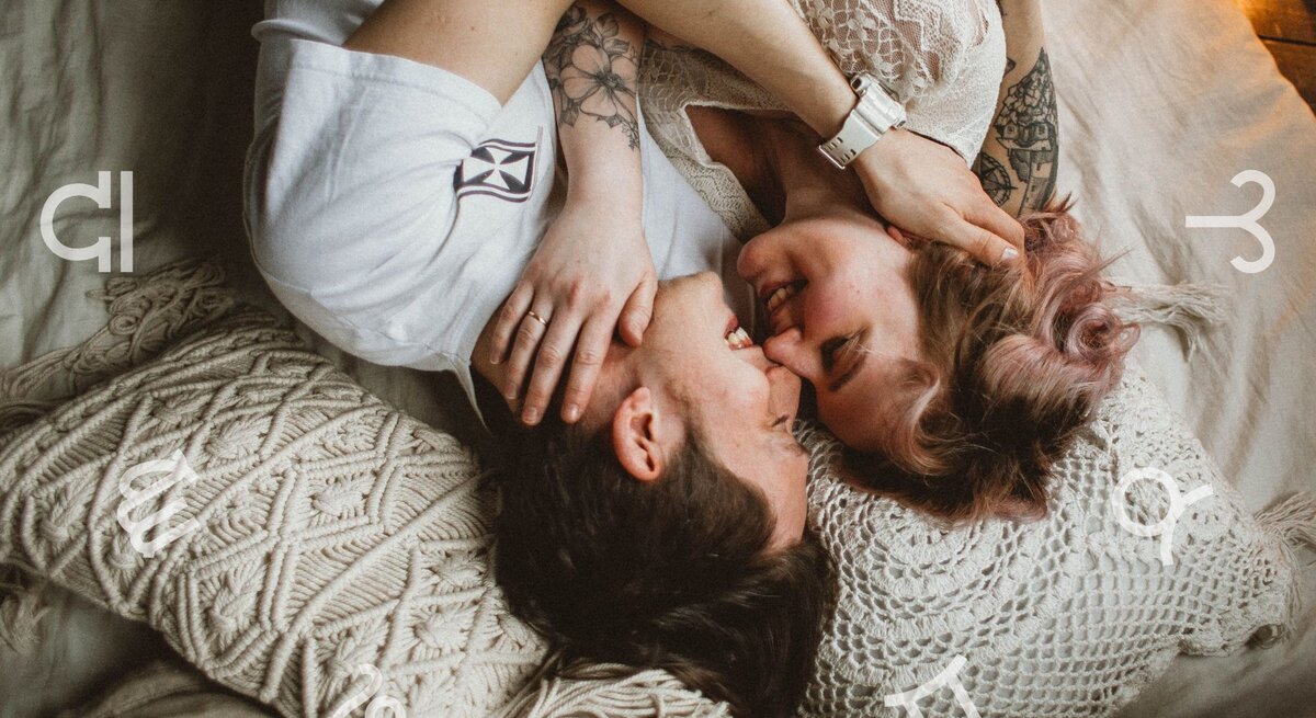 Ein verliebtes Paar liegt eng umschlungen auf einem Bett mit gemütlichen, gehäkelten Kissen. Beide lächeln sich an, während um sie herum astrologische Symbole der Tierkreiszeichen schweben. Die Szene strahlt Intimität, Romantik und eine harmonische Atmosphäre aus.