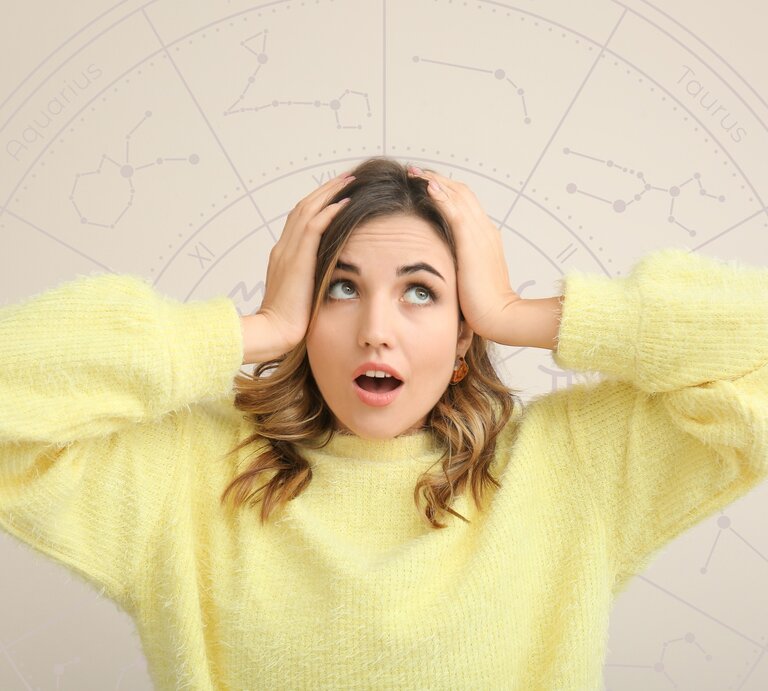 Frau in gelbem Pullover schaut überrascht und hält sich die Hände an den Kopf. Im Hintergrund ist ein astrologisches Rad mit Tierkreiszeichen wie Aquarius, Taurus und Gemini zu sehen. | © WinWin.artlab und pixelshot via Canva.com [M]
