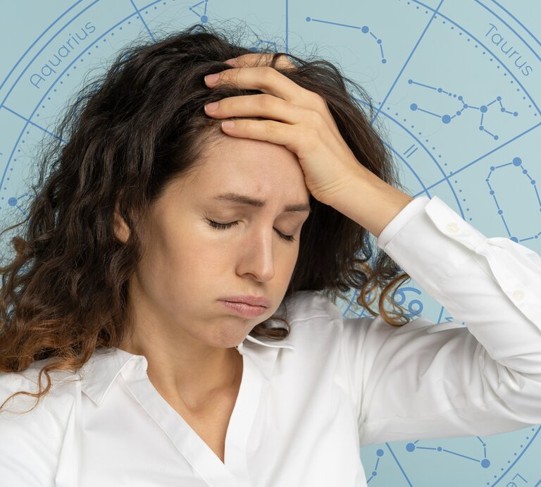 Eine Frau mit lockigem Haar und weißer Bluse hält sich mit geschlossenen Augen die Hand an die Stirn und wirkt erschöpft oder nachdenklich. Im Hintergrund ist ein astrologisches Rad mit den Sternzeichen zu sehen, was auf einen Zusammenhang zwischen ihrem Gemütszustand und astrologischen Einflüssen hinweist. | © WinWin.artlab und dimaberlinphotos via Canva.com [M]
