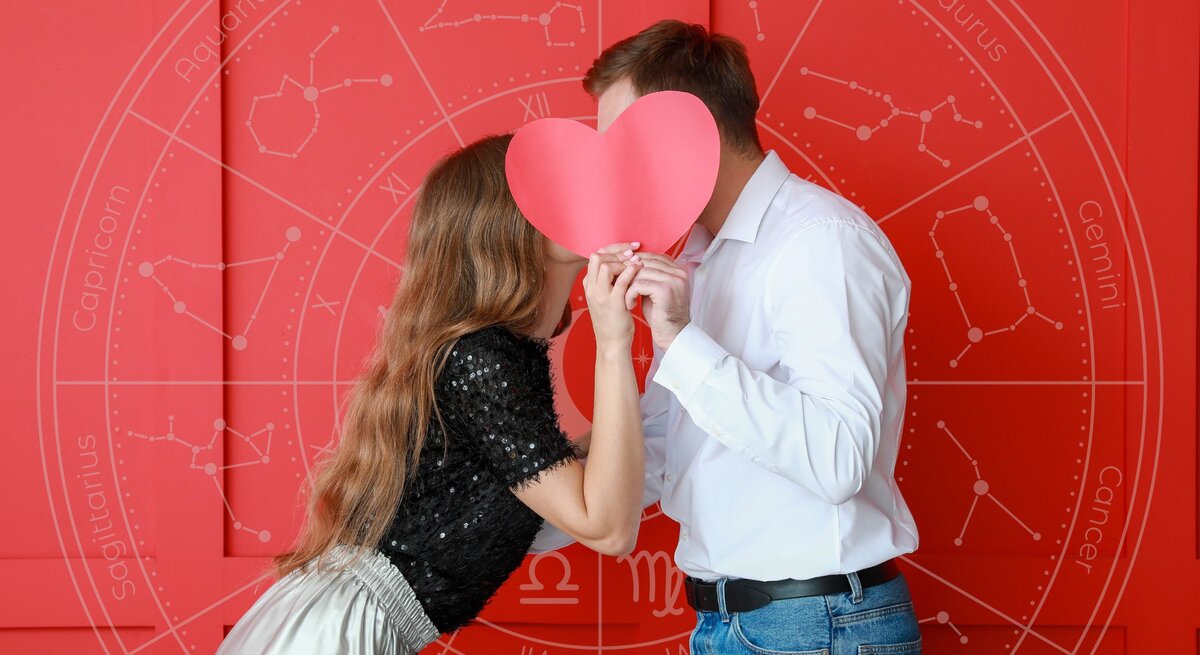 Ein Paar steht vor einem roten Hintergrund und hält ein großes, rotes Papierherz vor ihre Gesichter, sodass sie dahinter verborgen sind, als ob sie sich küssen würden. Im Hintergrund ist ein astrologisches Rad mit den Sternzeichen zu sehen, was eine Verbindung zwischen Liebe und Astrologie darstellt.