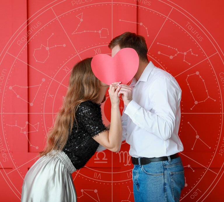 Ein Paar steht vor einem roten Hintergrund und hält ein großes, rotes Papierherz vor ihre Gesichter, sodass sie dahinter verborgen sind, als ob sie sich küssen würden. Im Hintergrund ist ein astrologisches Rad mit den Sternzeichen zu sehen, was eine Verbindung zwischen Liebe und Astrologie darstellt. | © WinWin.artlab und pixelshot via Canva.com [M]