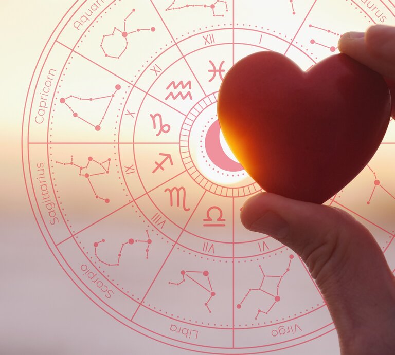 Eine Hand hält ein herzförmiges Objekt vor einem astrologischen Rad, das die Sternzeichen und ihre Symbole zeigt. Im Hintergrund geht die Sonne auf oder unter, und das Licht scheint durch das Herz. Das Bild symbolisiert die Verbindung von Liebe und Astrologie. | © Getty Images Pro/ snowflock und WinWin.artlab via Canva.com [M]