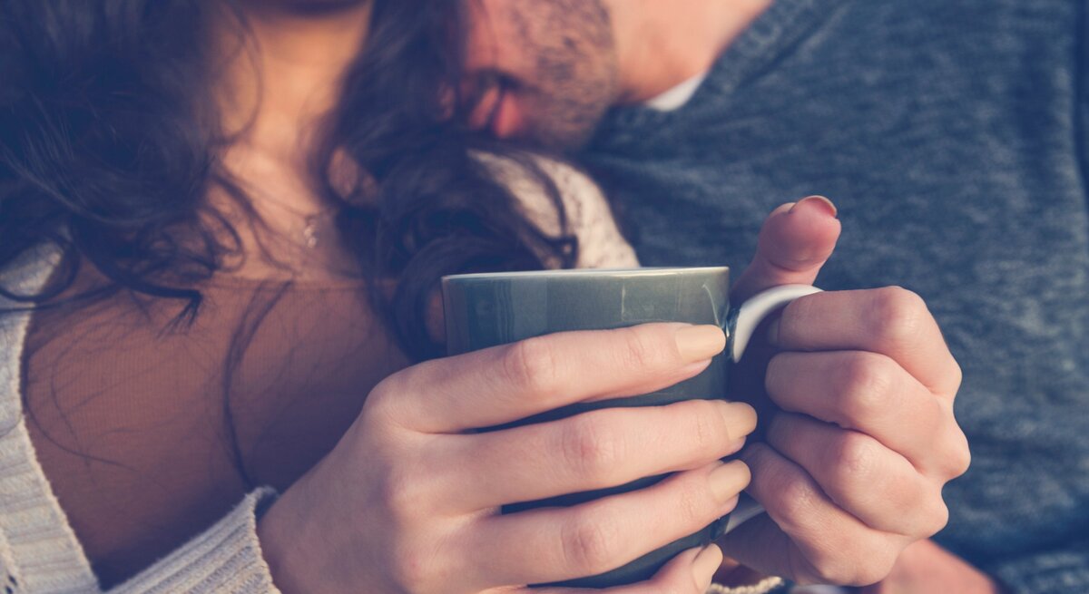 Ein Paar sitzt nah beieinander, während die Frau eine Tasse in beiden Händen hält. Der Mann lehnt seinen Kopf sanft an ihre Schulter, und beide wirken entspannt und verbunden. Die Frau lächelt leicht, während sie die Wärme der Tasse genießt. Das Bild vermittelt eine gemütliche, intime Atmosphäre und die Vertrautheit zwischen den beiden.
