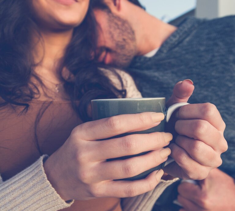 Ein Paar sitzt nah beieinander, während die Frau eine Tasse in beiden Händen hält. Der Mann lehnt seinen Kopf sanft an ihre Schulter, und beide wirken entspannt und verbunden. Die Frau lächelt leicht, während sie die Wärme der Tasse genießt. Das Bild vermittelt eine gemütliche, intime Atmosphäre und die Vertrautheit zwischen den beiden. | © Getty Images Signature/ Courtney Keating via Canva.com