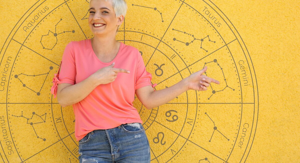 Eine lächelnde Frau mit kurzem, blondem Haar steht vor einer gelben Wand, auf der ein astrologisches Rad mit den Tierkreiszeichen abgebildet ist. Sie trägt ein pinkfarbenes T-Shirt und blaue Jeans und zeigt mit beiden Händen auf den Bereich des Horoskops. Die Szene wirkt freundlich und einladend, mit einem sonnigen Hintergrund, der die fröhliche Stimmung unterstreicht.