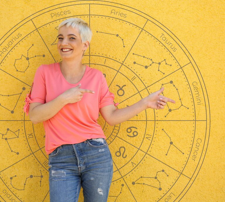Eine lächelnde Frau mit kurzem, blondem Haar steht vor einer gelben Wand, auf der ein astrologisches Rad mit den Tierkreiszeichen abgebildet ist. Sie trägt ein pinkfarbenes T-Shirt und blaue Jeans und zeigt mit beiden Händen auf den Bereich des Horoskops. Die Szene wirkt freundlich und einladend, mit einem sonnigen Hintergrund, der die fröhliche Stimmung unterstreicht. | © Coloures-Pic und WinWin.artlab via Canva.com [M]