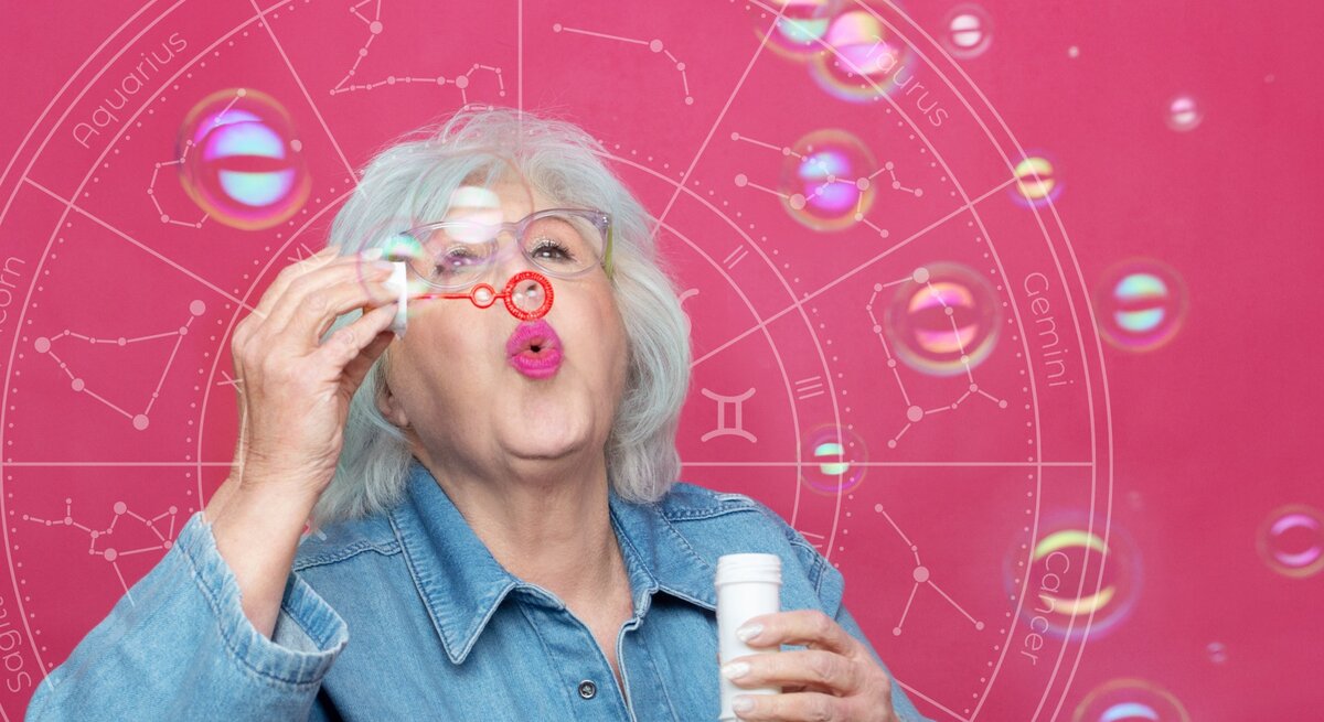 Eine ältere Frau mit weißem Haar und einer Brille bläst Seifenblasen in die Luft. Sie trägt eine Jeansjacke und steht vor einem pinkfarbenen Hintergrund, auf dem ein astrologisches Rad mit den Tierkreiszeichen angedeutet ist. Die Seifenblasen schweben vor ihr und reflektieren bunte Lichtpunkte, die eine spielerische und leichte Atmosphäre erzeugen.
