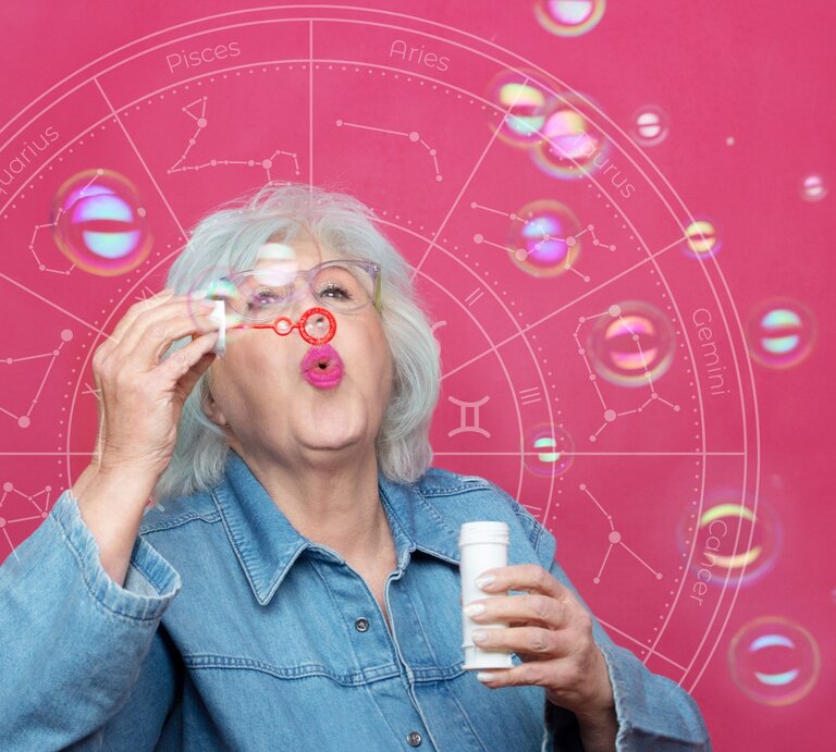 Eine ältere Frau mit weißem Haar und einer Brille bläst Seifenblasen in die Luft. Sie trägt eine Jeansjacke und steht vor einem pinkfarbenen Hintergrund, auf dem ein astrologisches Rad mit den Tierkreiszeichen angedeutet ist. Die Seifenblasen schweben vor ihr und reflektieren bunte Lichtpunkte, die eine spielerische und leichte Atmosphäre erzeugen. | © Getty Images/ William DondyK und WinWin.artlab via Canva.com [M]
