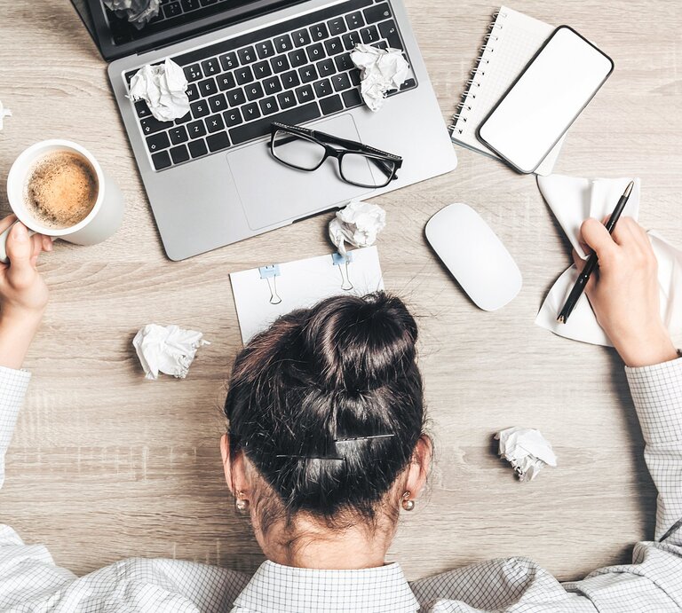 Eine Person liegt erschöpft mit dem Kopf auf einem Stapel von Formularen und Dokumenten, darüber ist die Tastatur eines Laptops zu sehen. Die Szene vermittelt Stress oder Überforderung durch Arbeit oder Bürokratie. | © oleksandrbedenyuk via Canva.com