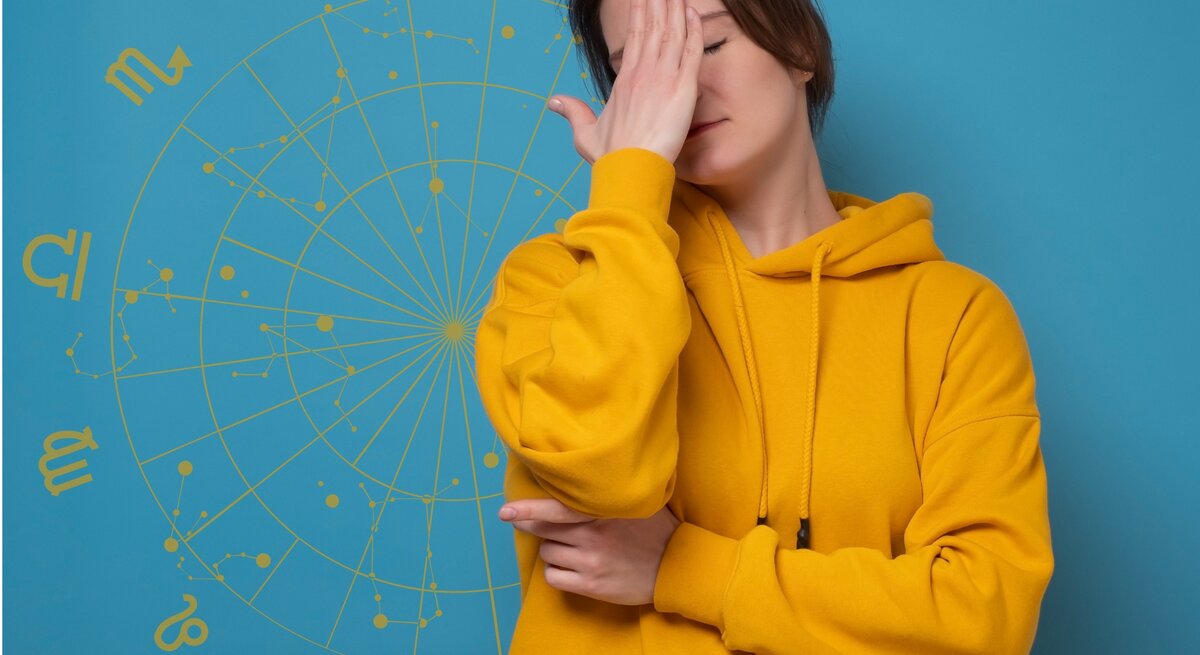 Eine Frau in einem gelben Hoodie steht vor einem blauen Hintergrund mit astrologischen Symbolen und hält sich frustriert die Hand vor das Gesicht. Ihre Haltung deutet auf Enttäuschung oder Erschöpfung hin, während das astrologische Rad im Hintergrund eine Verbindung zu Horoskopen oder astrologischen Themen andeutet.