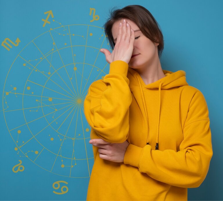 Eine Frau in einem gelben Hoodie steht vor einem blauen Hintergrund mit astrologischen Symbolen und hält sich frustriert die Hand vor das Gesicht. Ihre Haltung deutet auf Enttäuschung oder Erschöpfung hin, während das astrologische Rad im Hintergrund eine Verbindung zu Horoskopen oder astrologischen Themen andeutet. | © Getty Images/ Koldunova_Anna, Gabrielle Scarlett und Chikovnaya via Canva.com [M]
