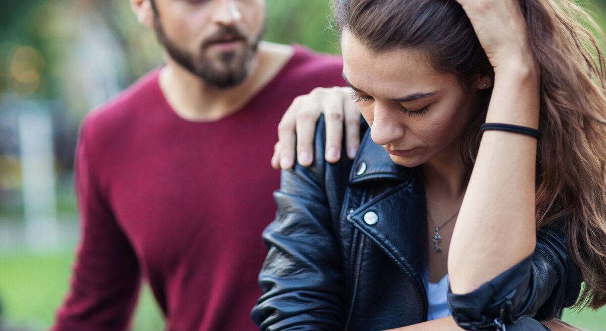 Ein Mann legt tröstend seine Hand auf die Schulter einer nachdenklich und traurig wirkenden Frau. Beide sitzen im Freien, die Frau hält ihren Kopf mit einer Hand und wirkt besorgt, während der Mann mitfühlend neben ihr sitzt.