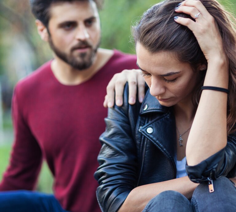 Ein Mann legt tröstend seine Hand auf die Schulter einer nachdenklich und traurig wirkenden Frau. Beide sitzen im Freien, die Frau hält ihren Kopf mit einer Hand und wirkt besorgt, während der Mann mitfühlend neben ihr sitzt. | © Getty Images Signature/ Mixmike