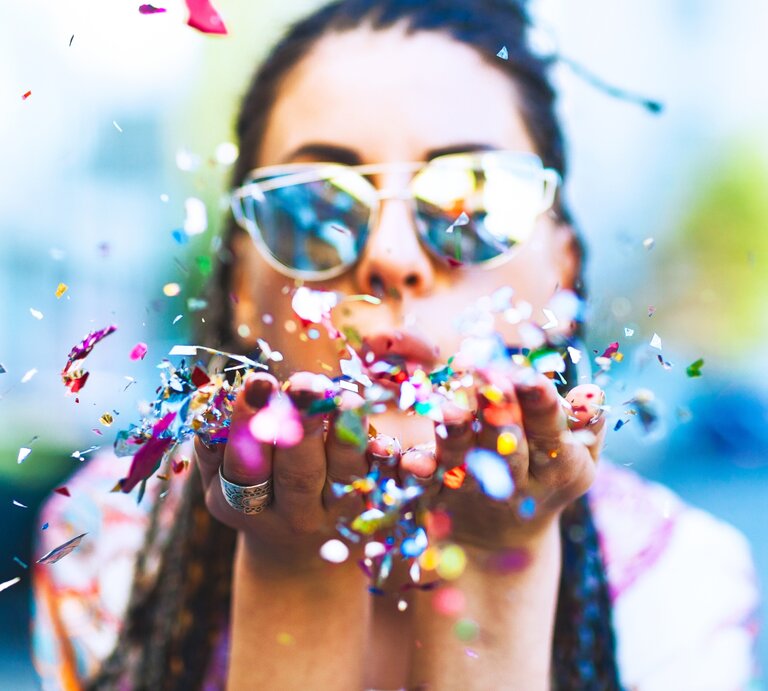 Eine Frau mit Sonnenbrille bläst farbenfrohe Konfettistücke in die Luft. Das Konfetti wirbelt um sie herum, während die lebendigen Farben eine fröhliche und festliche Atmosphäre erzeugen. | © Getty Images Signature/ Jasmina007