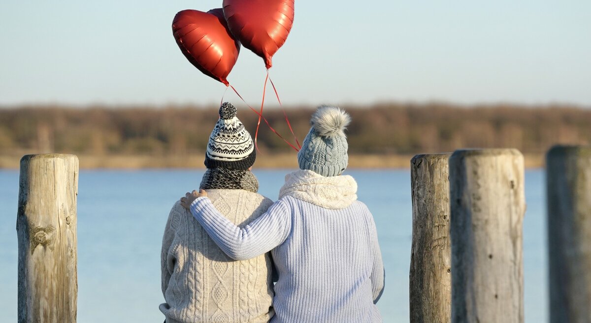 Zwei Personen sitzen eng umschlungen auf einem Holzsteg an einem See, mit Blick auf das Wasser. Sie tragen warme Winterkleidung und halten drei rote Herzballons in der Hand, die in die Luft steigen. Die Szene vermittelt eine ruhige und romantische Atmosphäre.