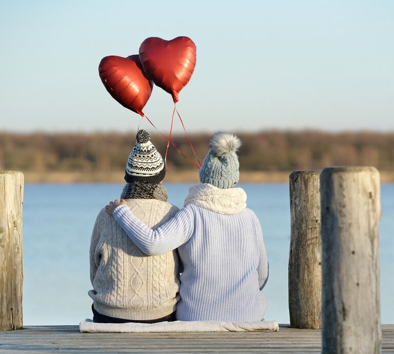 Zwei Personen sitzen eng umschlungen auf einem Holzsteg an einem See, mit Blick auf das Wasser. Sie tragen warme Winterkleidung und halten drei rote Herzballons in der Hand, die in die Luft steigen. Die Szene vermittelt eine ruhige und romantische Atmosphäre. | © Adobe Stock/ Jenny Sturm