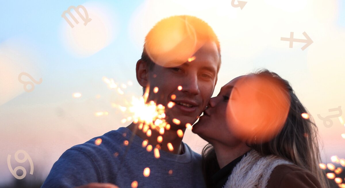 Das Bild zeigt ein Pärchen in einer romantischen Stimmung bei Sonnenuntergang. Die Frau gibt dem Mann einen Kuss auf die Wange, während er eine brennende Wunderkerze in der Hand hält. Im Hintergrund leuchten helle Lichtreflexe, die eine warme, festliche Atmosphäre erzeugen. Um die beiden herum sind Symbole der Sternzeichen wie Waage, Löwe und Skorpion zu sehen, was auf ein astrologisches Thema hinweist. Die Szene wirkt intim und fröhlich und vermittelt eine stimmungsvolle, romantische Stimmung.