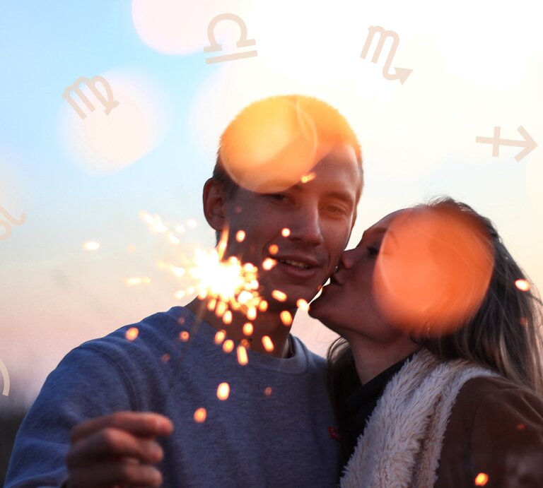 Das Bild zeigt ein Pärchen in einer romantischen Stimmung bei Sonnenuntergang. Die Frau gibt dem Mann einen Kuss auf die Wange, während er eine brennende Wunderkerze in der Hand hält. Im Hintergrund leuchten helle Lichtreflexe, die eine warme, festliche Atmosphäre erzeugen. Um die beiden herum sind Symbole der Sternzeichen wie Waage, Löwe und Skorpion zu sehen, was auf ein astrologisches Thema hinweist. Die Szene wirkt intim und fröhlich und vermittelt eine stimmungsvolle, romantische Stimmung. | © Getty Images/ Elchinm und Chikovnaya via Canva.com [M]