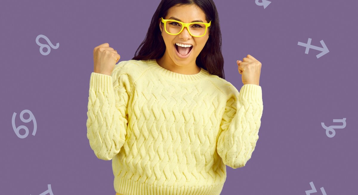 Eine fröhliche Frau mit langen, dunklen Haaren, die gelbe Brille und einen gelben Pullover trägt, zeigt freudig beide Fäuste. Im Hintergrund sind astrologische Symbole auf einem lilafarbenen Hintergrund verteilt.