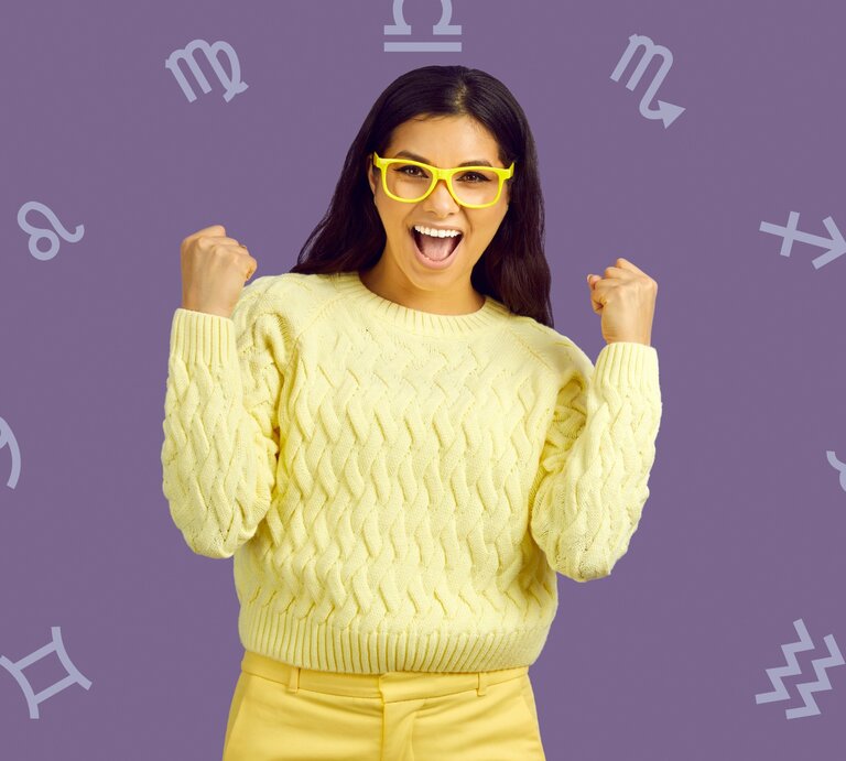 Eine fröhliche Frau mit langen, dunklen Haaren, die gelbe Brille und einen gelben Pullover trägt, zeigt freudig beide Fäuste. Im Hintergrund sind astrologische Symbole auf einem lilafarbenen Hintergrund verteilt. | © Chikovnaya und studioroman via Canva.com [M]
