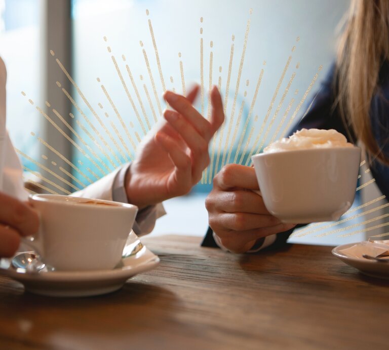 Zwei Personen sitzen in einem Café und halten Tassen mit Kaffee in den Händen, während sie sich unterhalten. Ihre Gesten und die Strahlen, die das Bild zieren, vermitteln eine energetische und positive Stimmung. | © Getty Images Signature/ webphotographeer und Julia Dreams via Canva.com [M].