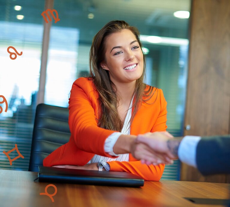 Eine lächelnde Frau in einem orangefarbenen Blazer sitzt in einem modernen Büro und schüttelt die Hand einer anderen Person, die teilweise im Bild sichtbar ist. Im Hintergrund schweben astrologische Symbole wie Löwe, Jungfrau, Krebs und Zwillinge in der Luft. | © Getty Images Signature/ sturti und Chikovnaya via Canva.com