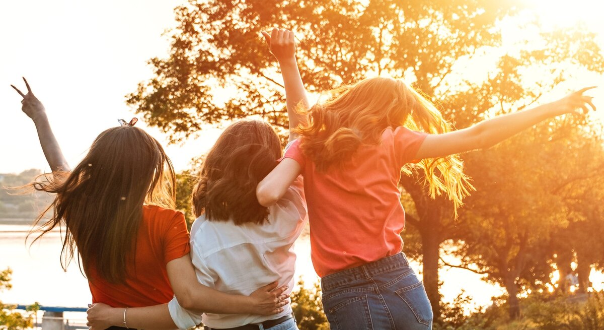 Drei junge Frauen stehen Arm in Arm mit dem Rücken zur Kamera, während sie die Arme freudig in die Luft werfen. Sie stehen in einer sonnigen Landschaft, umgeben von Bäumen, mit Blick auf einen Fluss oder See im Hintergrund. Die Sonne scheint warm und strahlend, was eine lebendige und fröhliche Atmosphäre schafft.