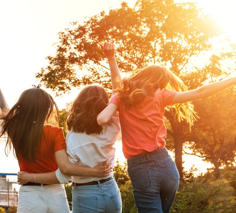 Drei junge Frauen stehen Arm in Arm mit dem Rücken zur Kamera, während sie die Arme freudig in die Luft werfen. Sie stehen in einer sonnigen Landschaft, umgeben von Bäumen, mit Blick auf einen Fluss oder See im Hintergrund. Die Sonne scheint warm und strahlend, was eine lebendige und fröhliche Atmosphäre schafft. | © irynakhabliuk via Canva.com