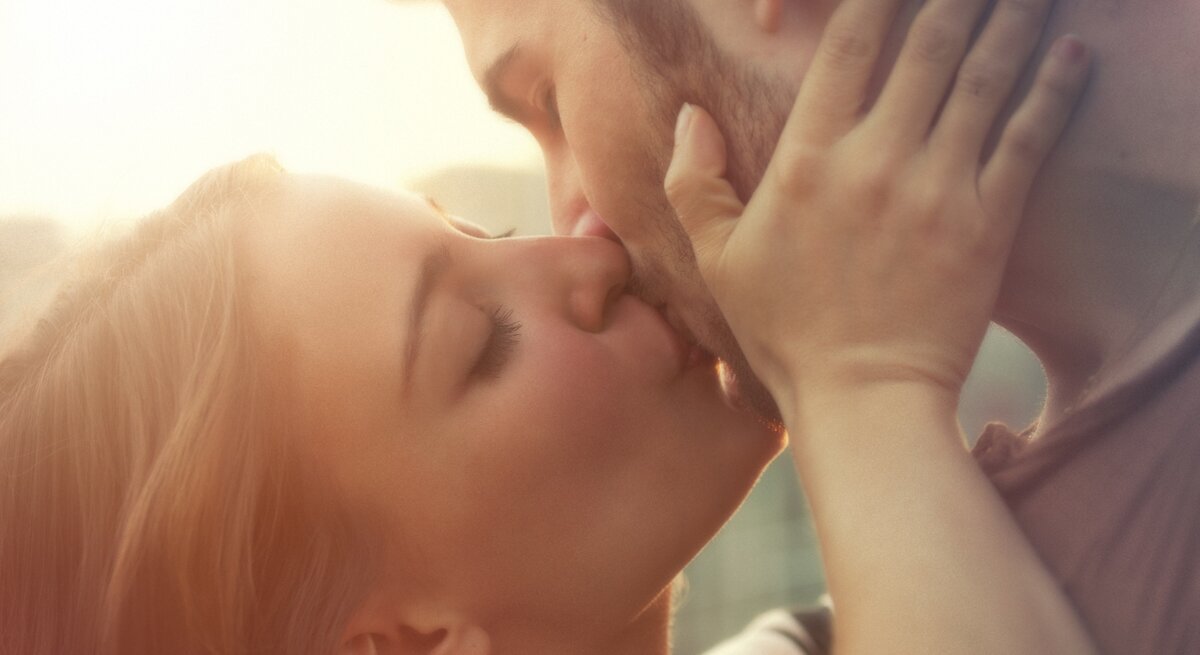 Ein Paar, bestehend aus einer Frau und einem Mann, teilt einen innigen Kuss. Die Frau hält das Gesicht des Mannes sanft mit beiden Händen, während die Sonne im Hintergrund für eine warme, romantische Beleuchtung sorgt.