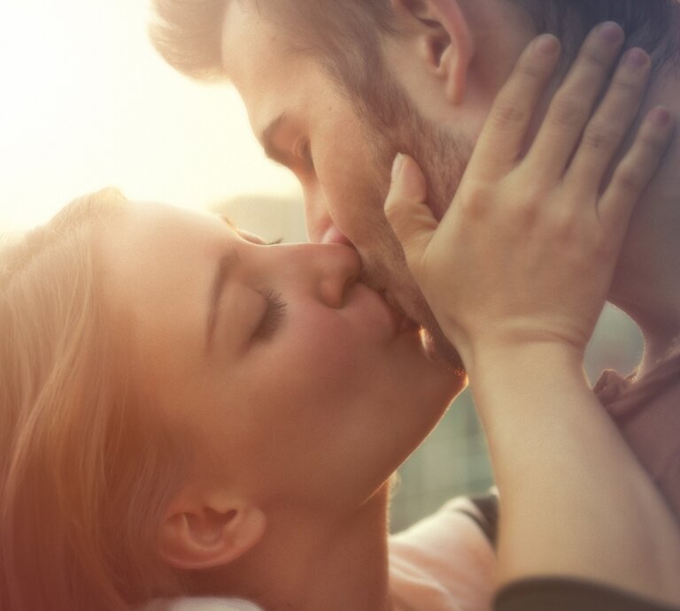Ein Paar, bestehend aus einer Frau und einem Mann, teilt einen innigen Kuss. Die Frau hält das Gesicht des Mannes sanft mit beiden Händen, während die Sonne im Hintergrund für eine warme, romantische Beleuchtung sorgt. | © Getty Images Signature/ mammuth