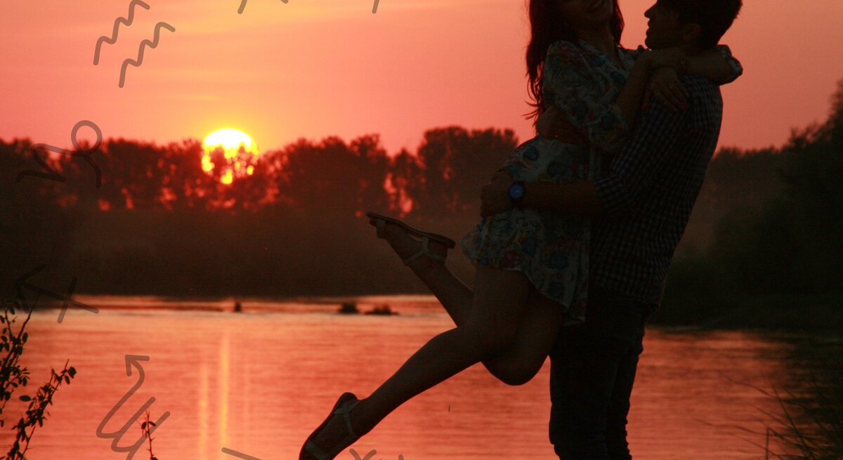 Ein Paar steht bei Sonnenuntergang am Ufer eines Flusses. Der Mann hebt die Frau lachend hoch, während sie beide glücklich in die Kamera blicken. Der Himmel ist in warmen Rottönen gefärbt, und die Reflexion der Sonne spiegelt sich im Wasser wider. Um das Paar herum schweben astrologische Symbole der Tierkreiszeichen, was eine Verbindung zur Astrologie und romantischen Themen andeutet. Die Szene vermittelt eine romantische und harmonische Atmosphäre.