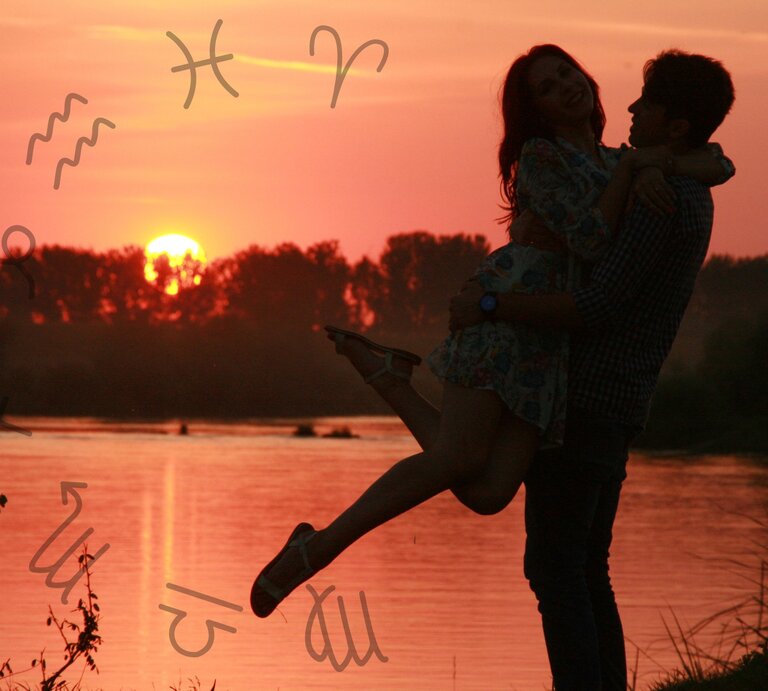 Ein Paar steht bei Sonnenuntergang am Ufer eines Flusses. Der Mann hebt die Frau lachend hoch, während sie beide glücklich in die Kamera blicken. Der Himmel ist in warmen Rottönen gefärbt, und die Reflexion der Sonne spiegelt sich im Wasser wider. Um das Paar herum schweben astrologische Symbole der Tierkreiszeichen, was eine Verbindung zur Astrologie und romantischen Themen andeutet. Die Szene vermittelt eine romantische und harmonische Atmosphäre. | © Keronyart's Images/ Be keronyart und pixabay/ AdinaVoicu via Canva.com [M]